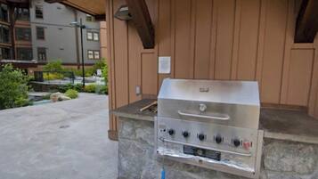 Una parrilla de acero inoxidable con cuatro quemadores, ubicada sobre una plataforma de piedra en un área exterior con una pérgola de madera.