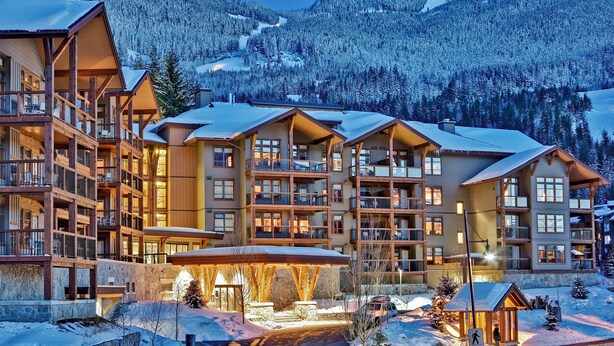 Un hôtel à plusieurs étages doté de balcons, d’un espace extérieur couvert et surplombant un paysage montagneux enneigé.