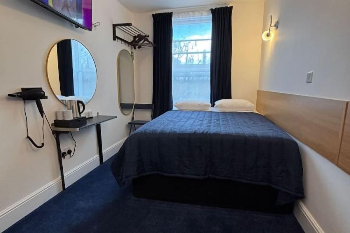 Quarto de hotel com cama, espelho redondo, secador de cabelo, porta-toalhas e janela com cortinas.