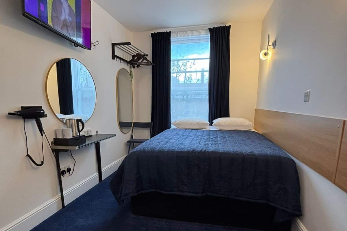 Habitación de hotel con cama, espejo, televisor, secador de pelo y ventana con cortinas.