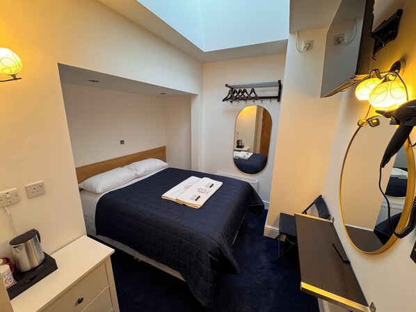 A hotel room with a bed, a bedside table with a kettle, a mirror, and a wall-mounted towel rack.