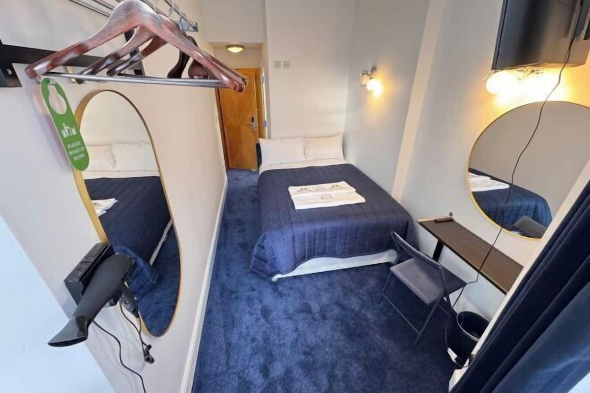 A small, compact room with a single bed, a desk, a chair, a mirror, and a coat rack.
