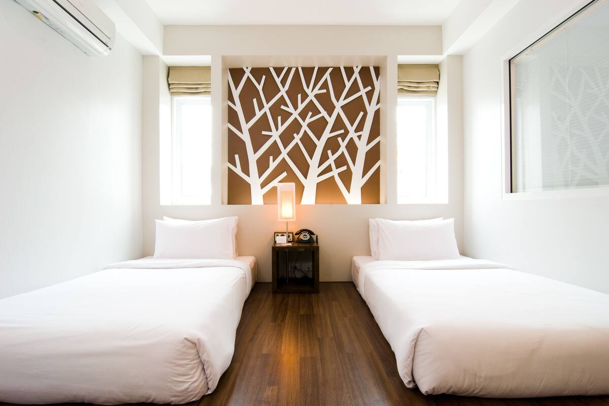 A hotel room with two beds, a wooden floor, a wall-mounted decorative panel, a bedside table with a lamp, and a window with curtains.
