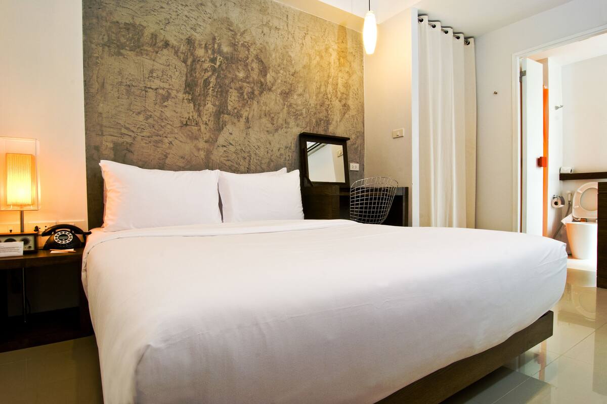 A large bed with white linens, a nightstand with a lamp and a telephone, a wall-mounted mirror, and a bathroom visible through an open door.