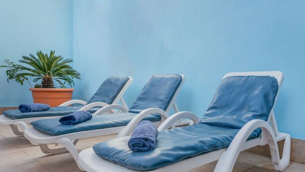 A row of white sun loungers with blue cushions and towels, against a light blue wall, with a potted plant to the left. - Qawra Point Holiday Complex (St. Paul's Bay)