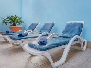 Une rangée de chaises longues blanches, agrémentées de coussins et de serviettes bleues, se dresse devant un mur bleu clair, avec une plante en pot placée à gauche.