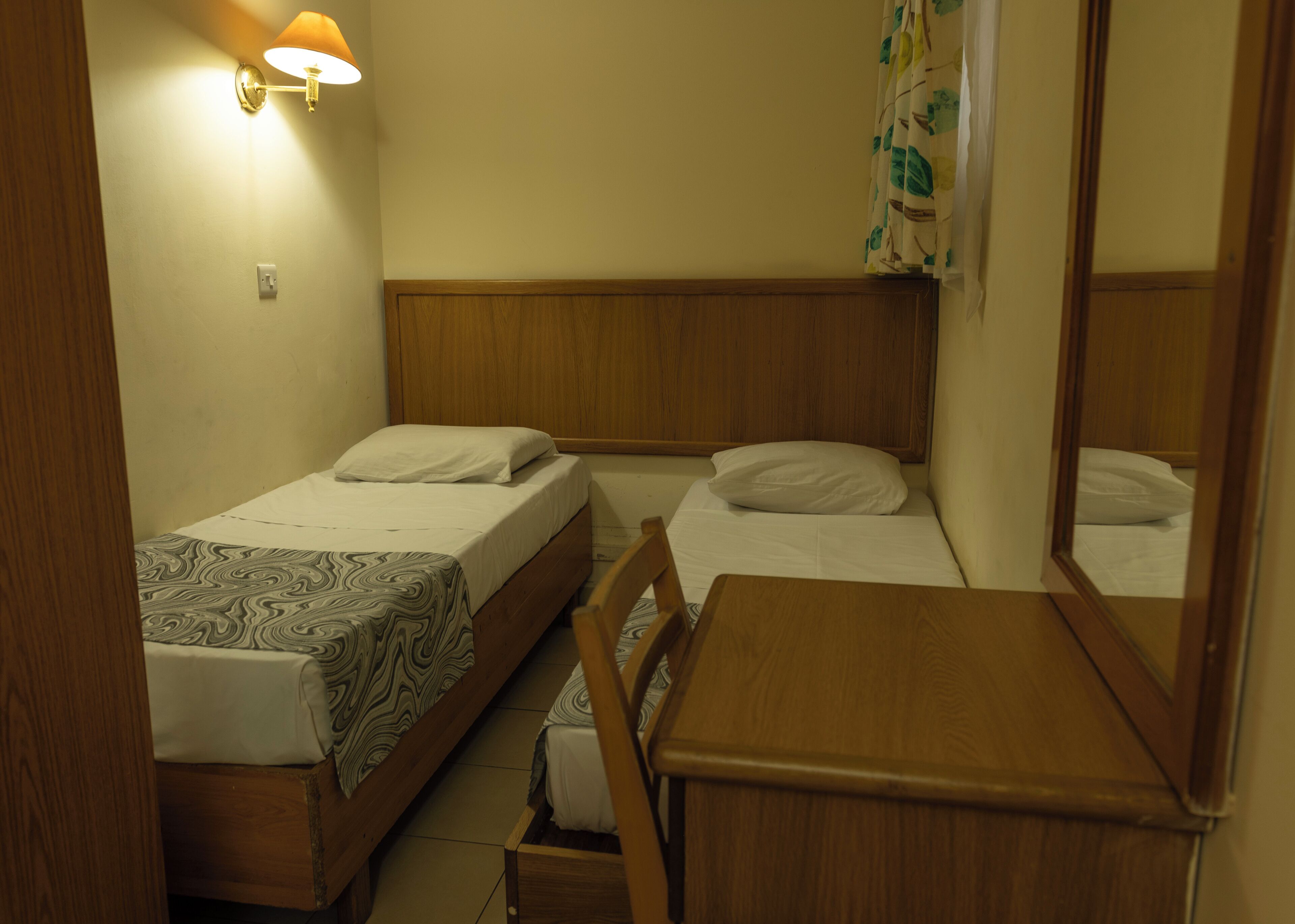 Een kleine, schone kamer met twee eenpersoonsbedden, een houten hoofdbord, een nachtkastje, een stoel en een wandlamp.