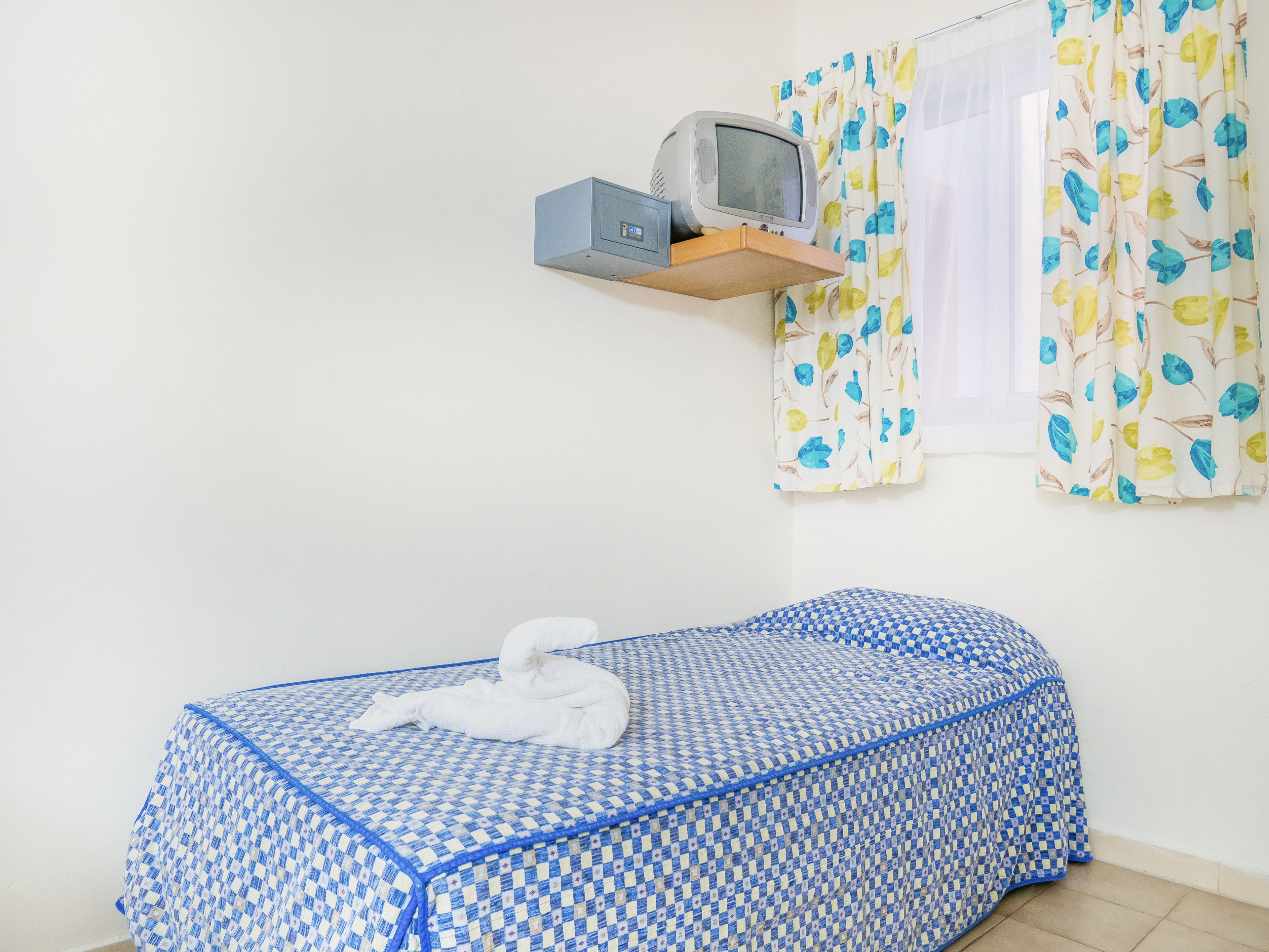 Una cama individual con una colcha azul y blanca a cuadros, un televisor pequeño sobre una estantería de madera y una ventana con cortinas estampadas.