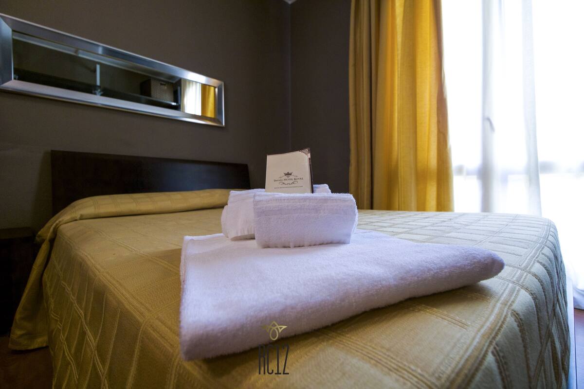 A hotel room with a neatly made bed, a headboard, a window with curtains, and a wall-mounted air conditioning unit.