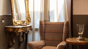 Una habitación con un espejo grande y ornamentado, un sillón a rayas, una mesita redonda y un ventanal con cortinas traslúcidas.