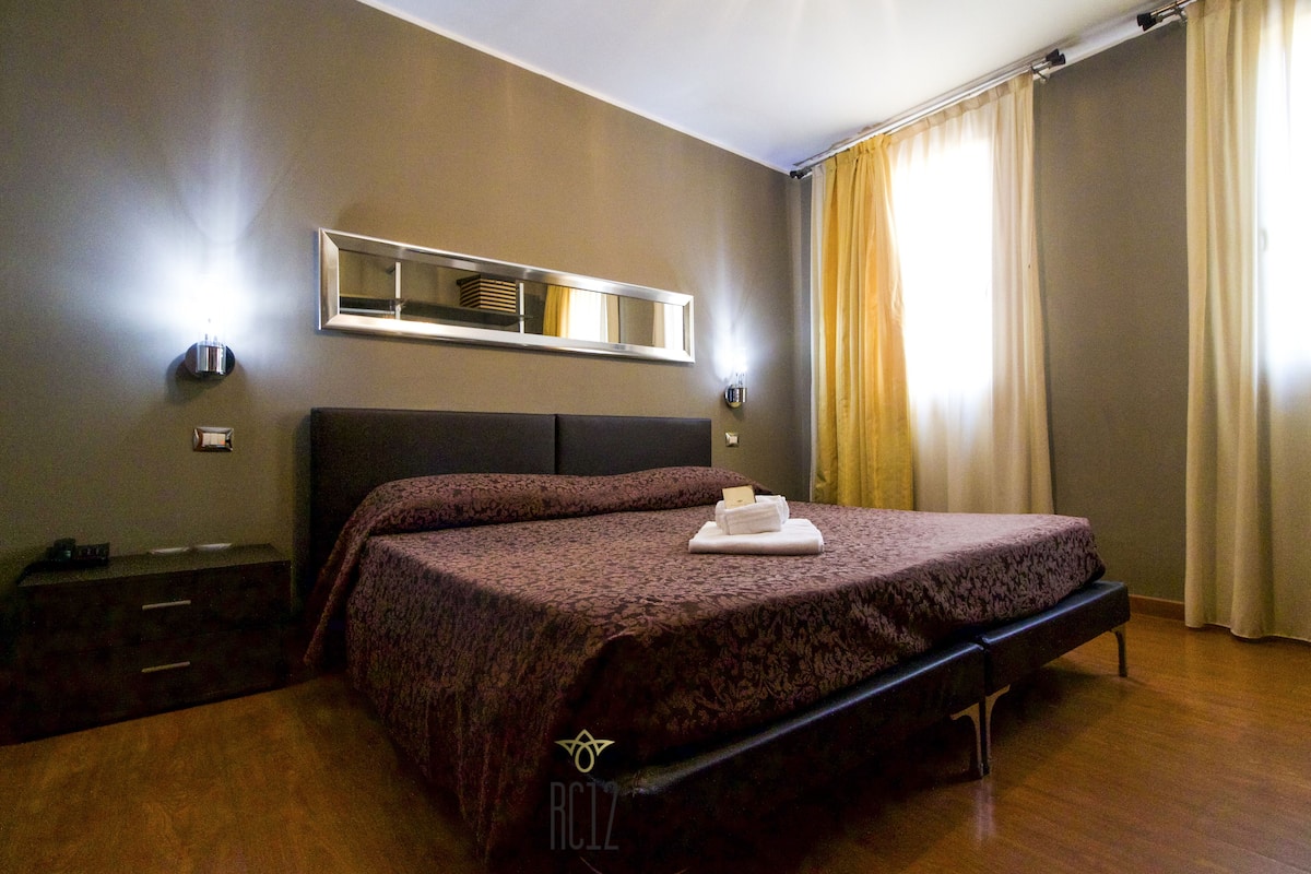 A hotel room with a large bed, a bedside table, a dresser, a mirror, and a window with curtains.