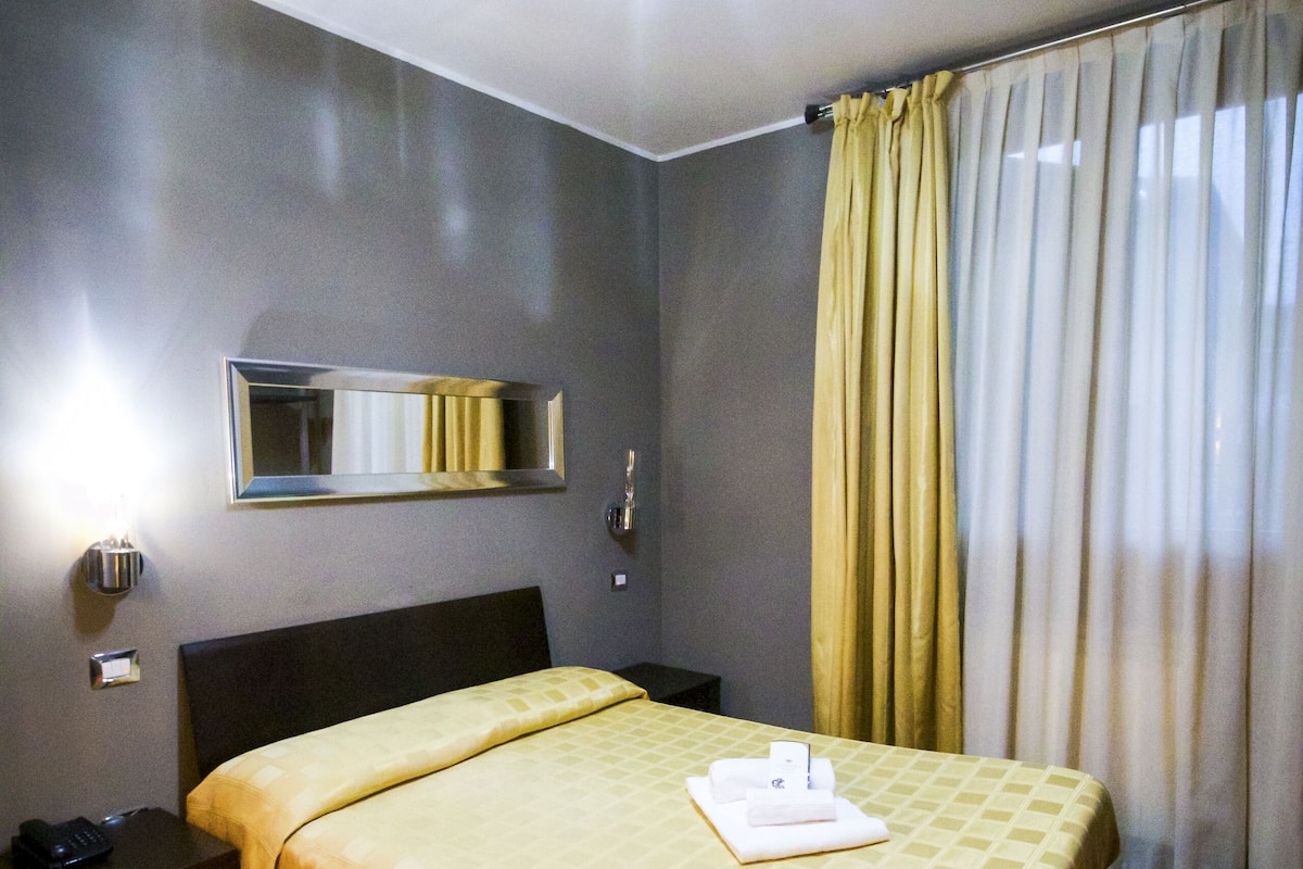 A hotel room with a bed, a bedside table, a mirror, and curtains.