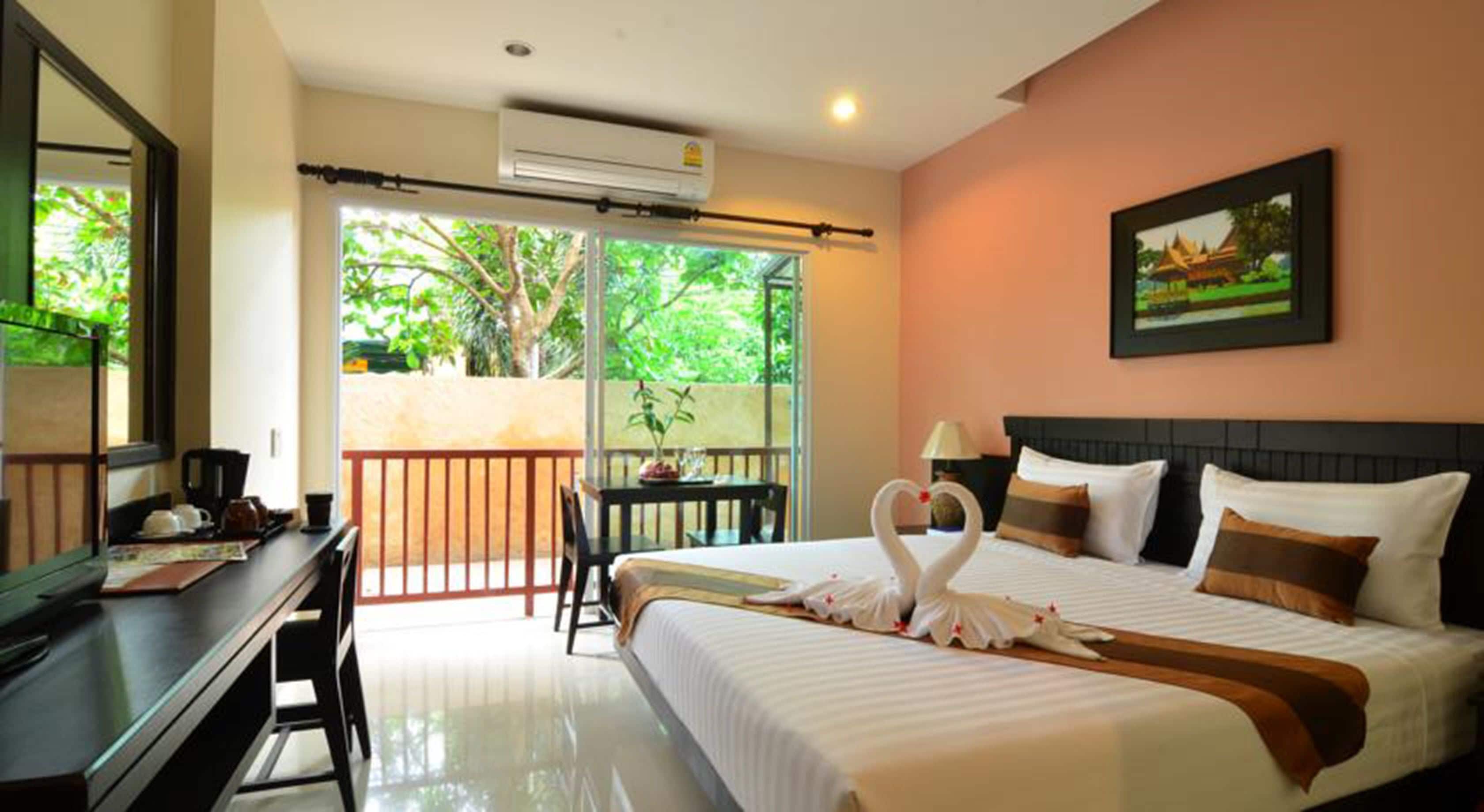 A hotel room with a bed, a desk, a chair, a balcony with a view of greenery, and a painting on the wall.