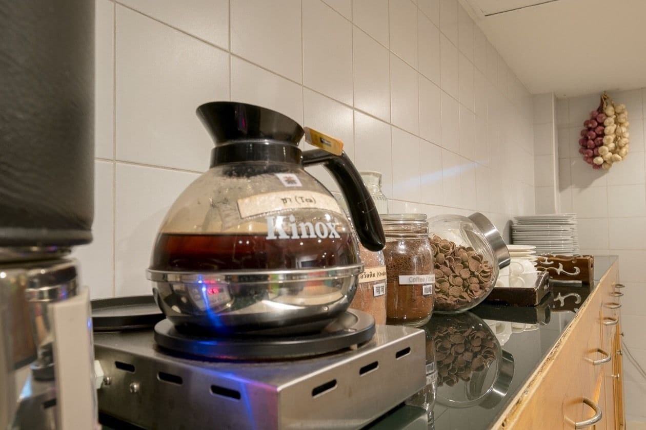 A kitchen with a coffee maker labeled "Kinox" on a stove, jars of coffee and chocolate, and a hanging bunch of garlic bulbs.