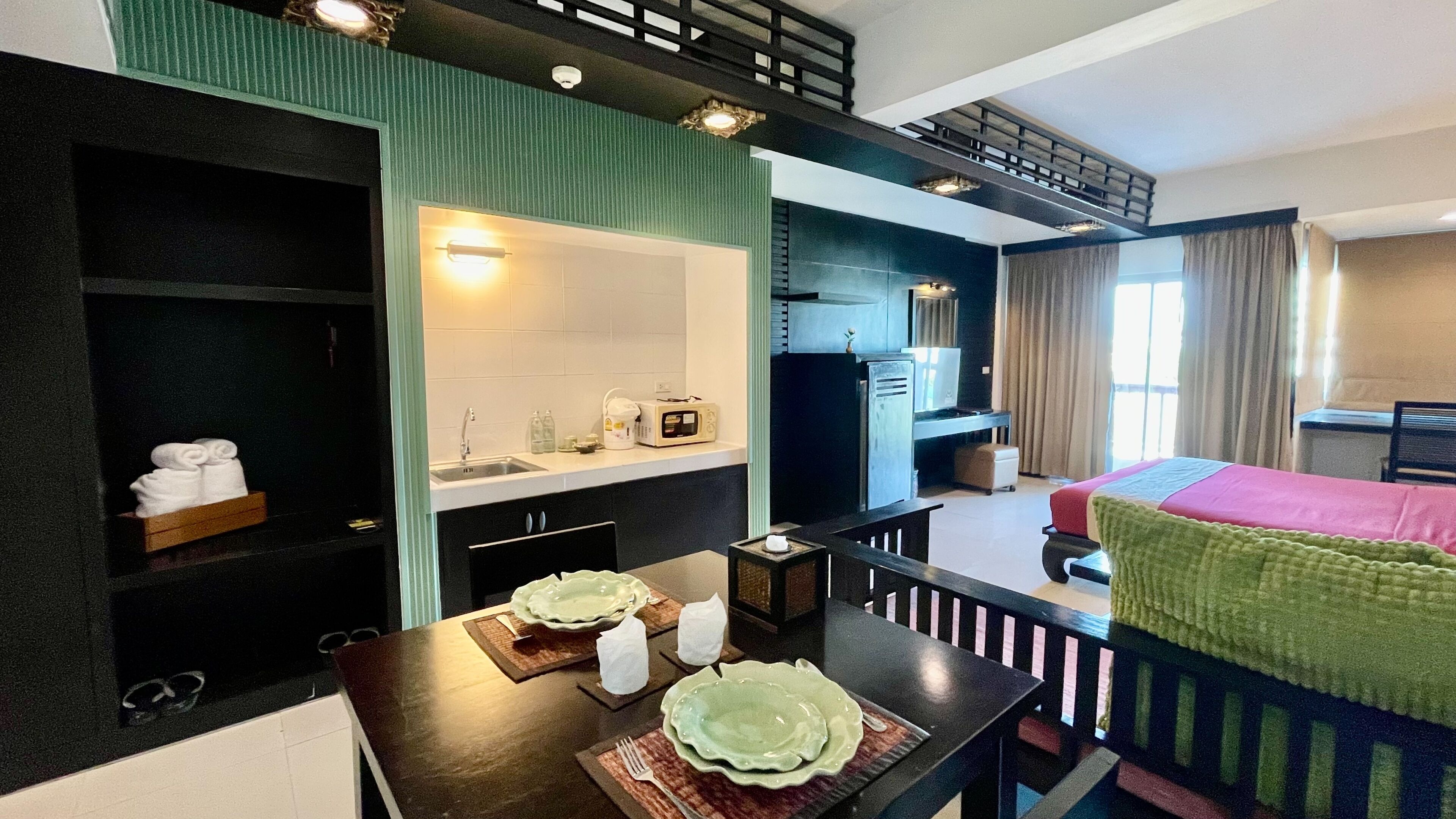 Une chambre d’hôtel avec une kitchenette, un lit recouvert d’une couverture verte, un coin repas avec une table dressée pour deux personnes, et une fenêtre avec des rideaux.