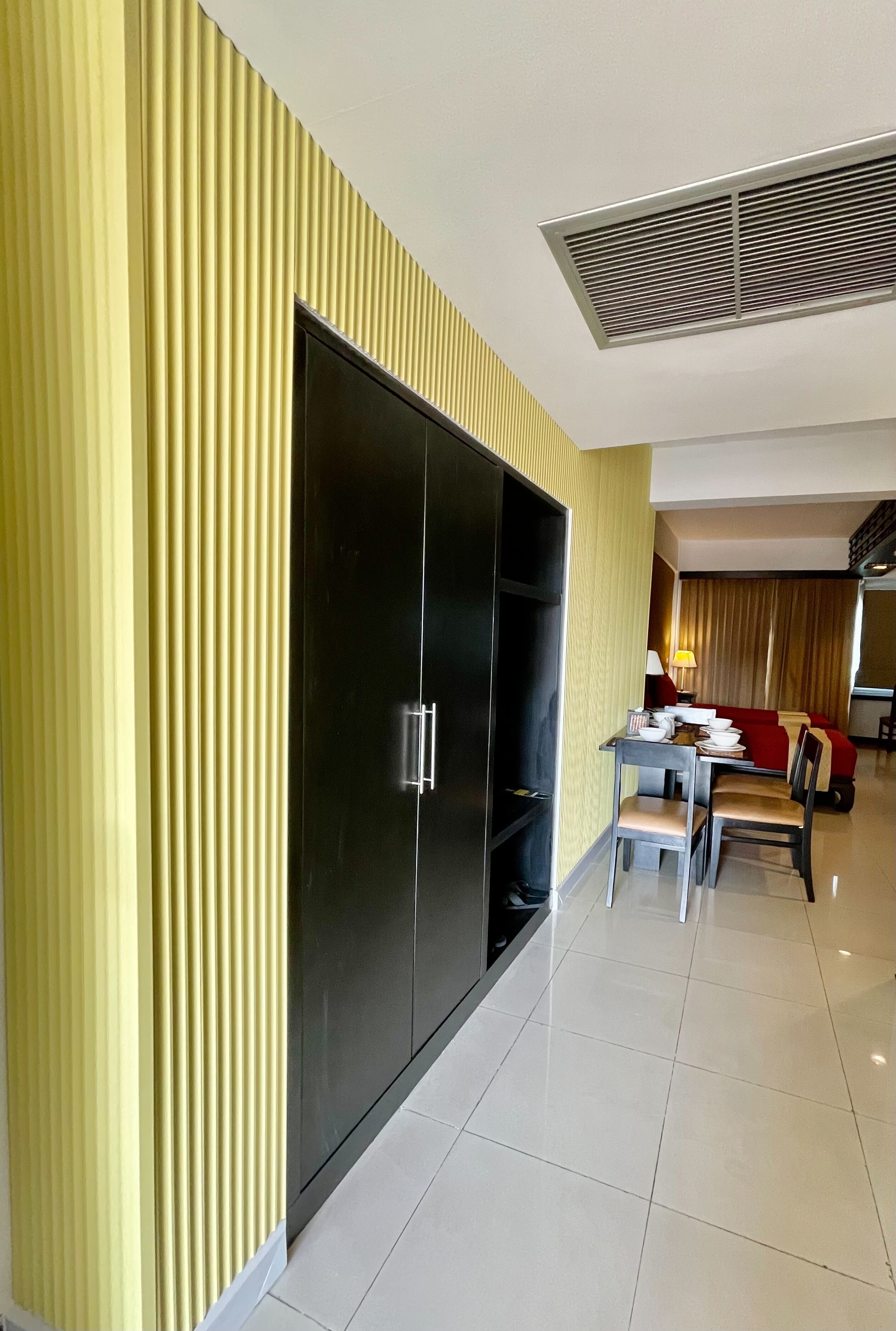 A modern hotel room with a large wardrobe, a dining area with a table and chairs, and a living area with a sofa and lamp.