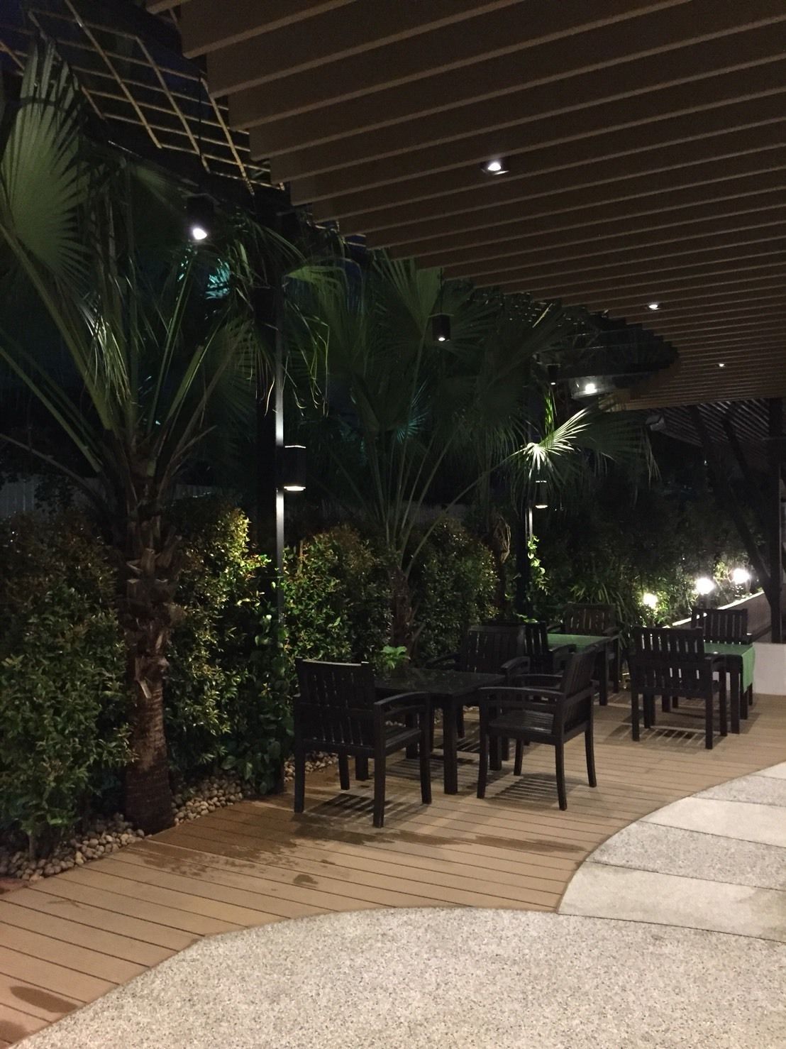An outdoor patio area with wooden flooring, surrounded by lush greenery including palm trees and shrubs. There are several tables and chairs arranged for dining or socializing.