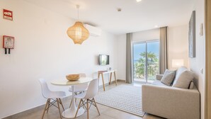 A modern living room with a sofa, a dining table with chairs, a TV, and a large window with a view of palm trees. - Hotel Clube Porto Mos (Lagos)