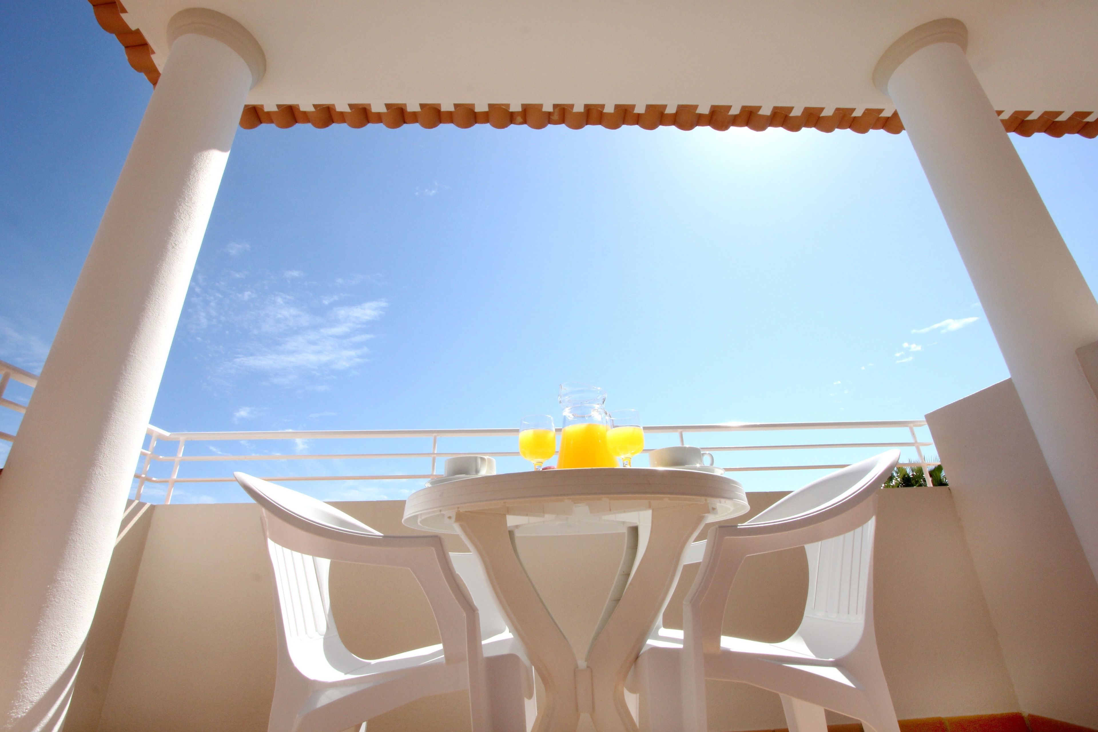 Un balcón con sillas y una mesa blancos, cielo azul despejado y un sol al fondo.