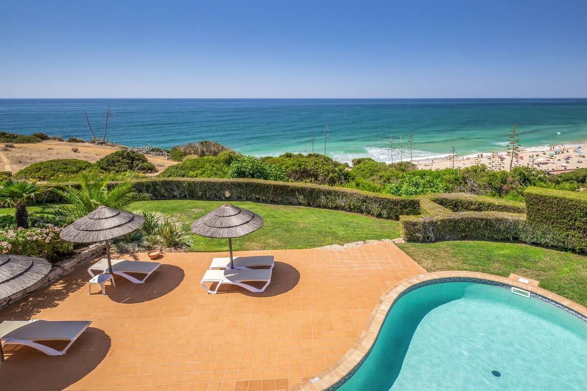 A pool area with lounge chairs and umbrellas, a well-maintained lawn, and a view of the beach and ocean.