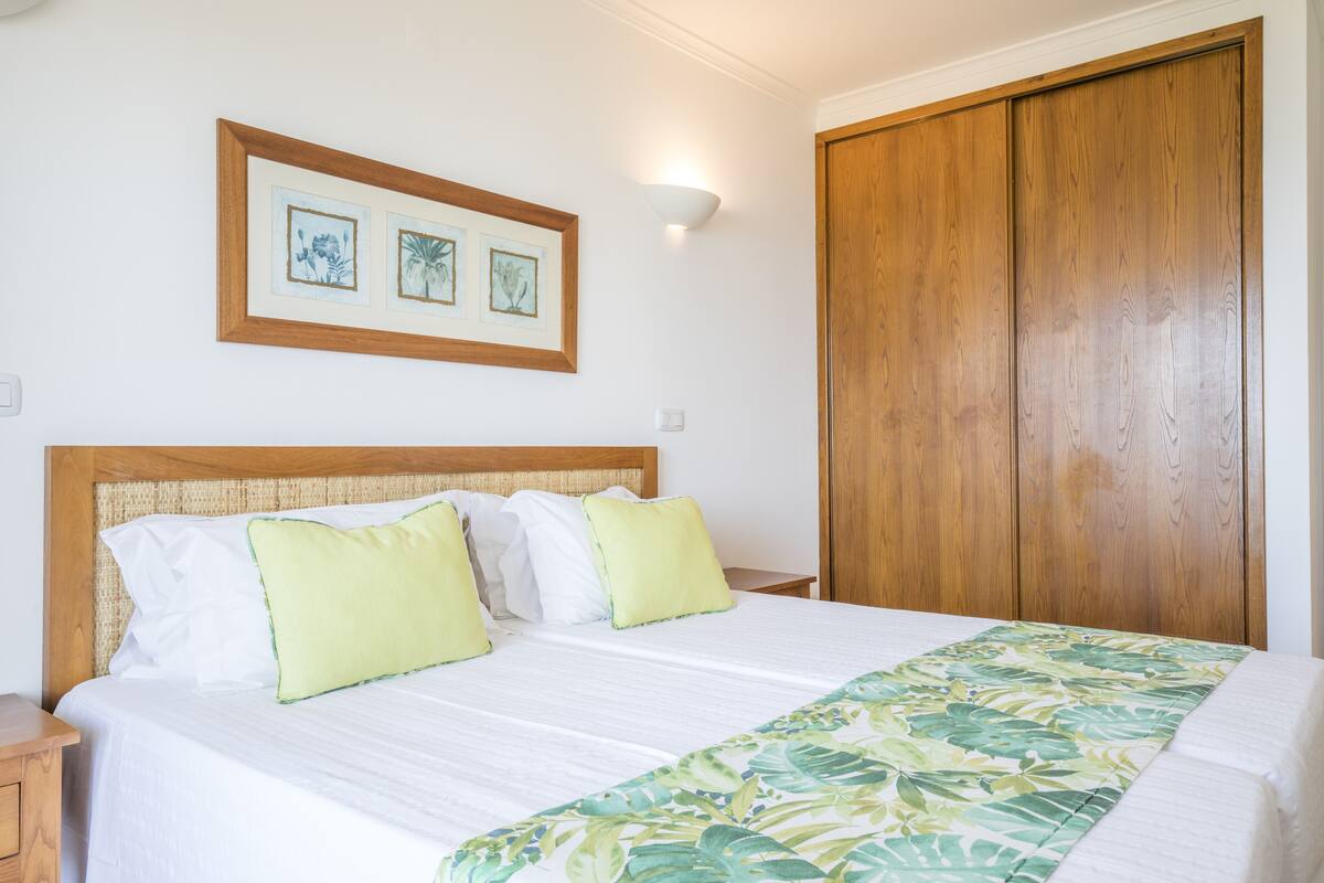 A neatly made bed with a floral bedspread, a wooden headboard, and two framed artworks above it.