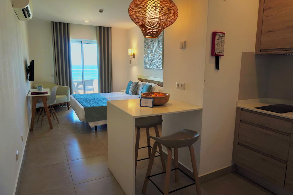 A compact hotel room with a bed, a small kitchenette, a dining area, and a view of the sea.