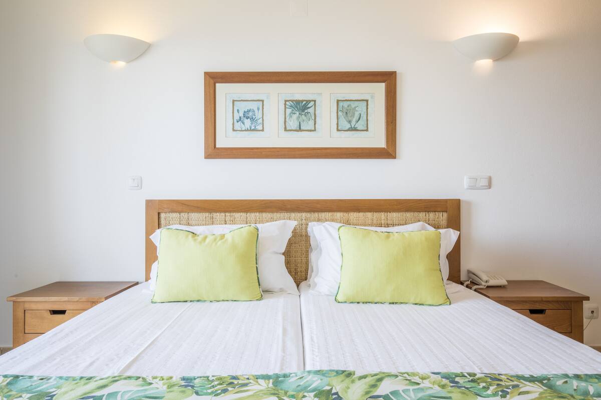 A neatly made bed with two pillows, a framed artwork above it, and a bedside table with a phone.