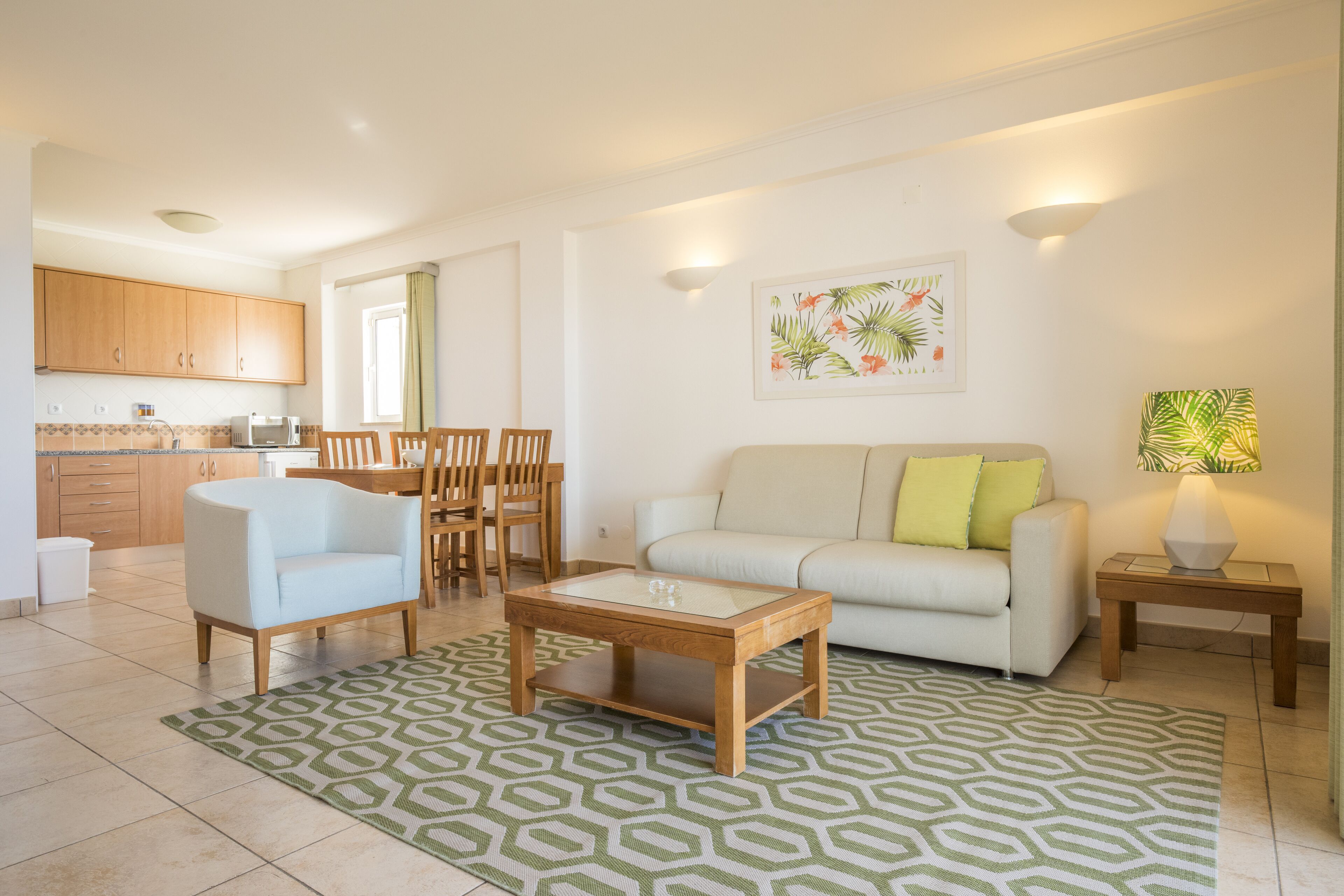 A living room with a sofa, coffee table, and chairs. There is a kitchen in the background with cabinets and a microwave.