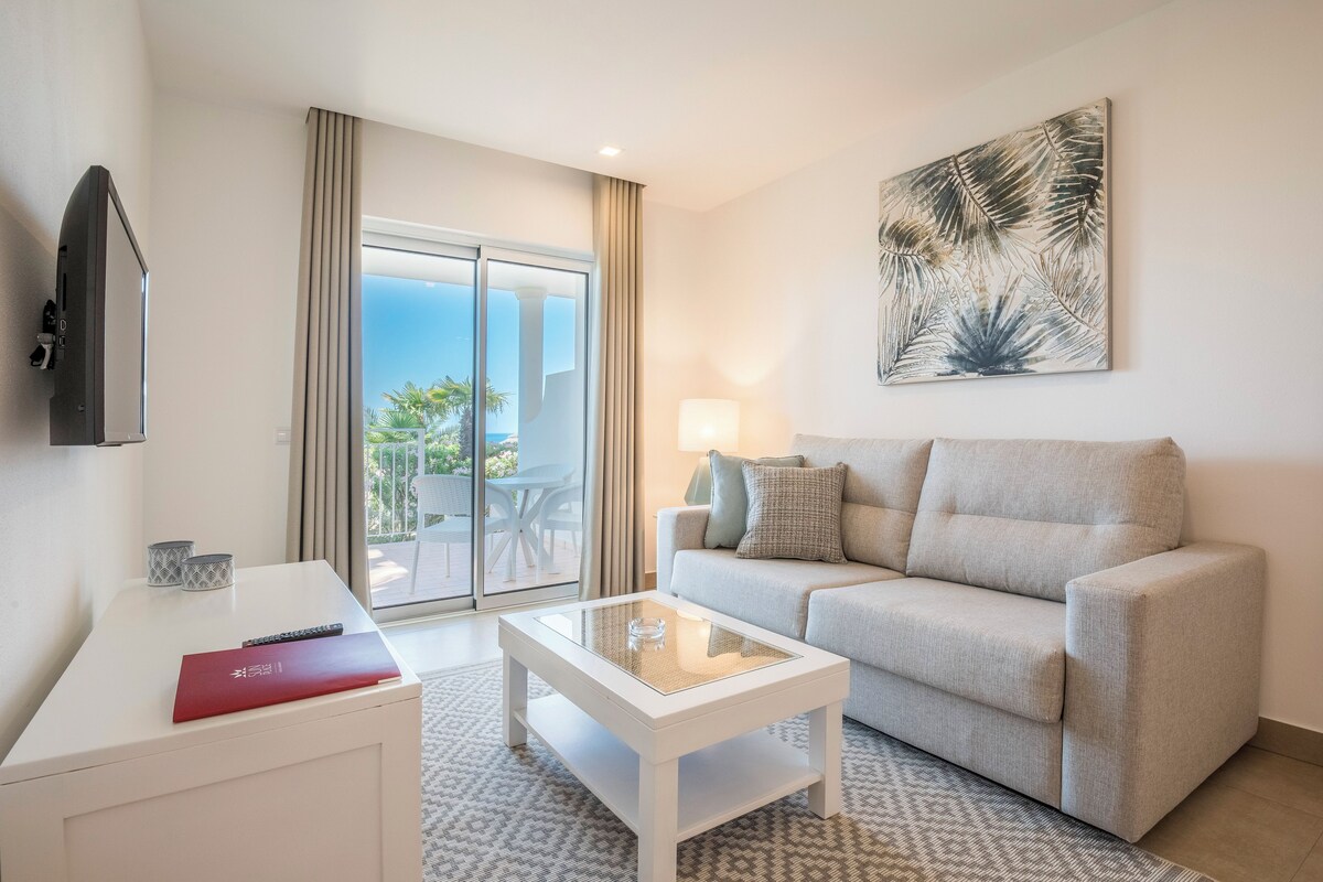 A modern hotel room with a sofa, a glass-top coffee table, a TV, and a view of the outdoors.
