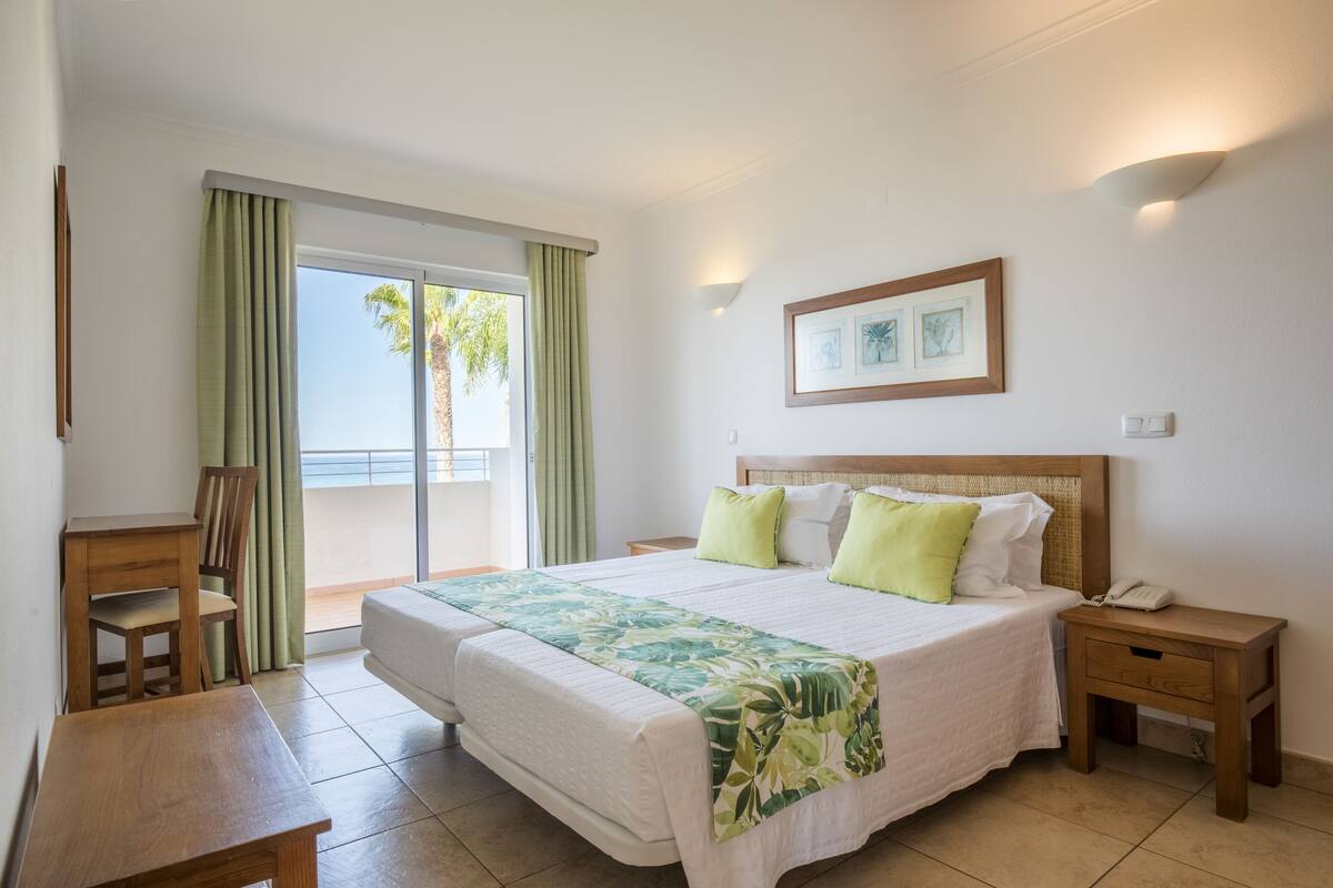 A hotel room with a bed, a wooden desk, a nightstand, a framed picture, and a view of palm trees through the window.