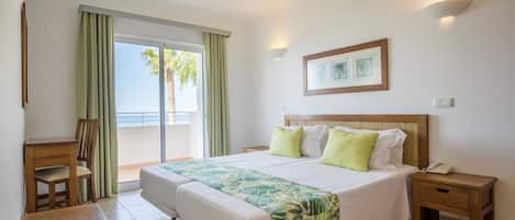 A hotel room with a bed, a wooden desk, a nightstand, a framed picture, and a view of palm trees through the window.