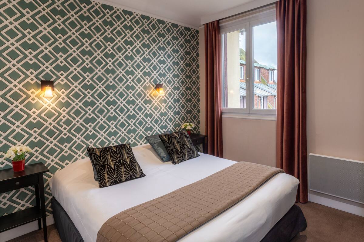 Een net opgemaakt bed met gekleurde kussens, een nachtkastje met een bloemenvaas, en een raam met gordijnen.