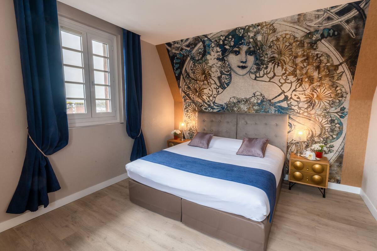 Een slaapkamer met een groot bed, een nachtkastje, wandtapijt en een raam met gordijnen.