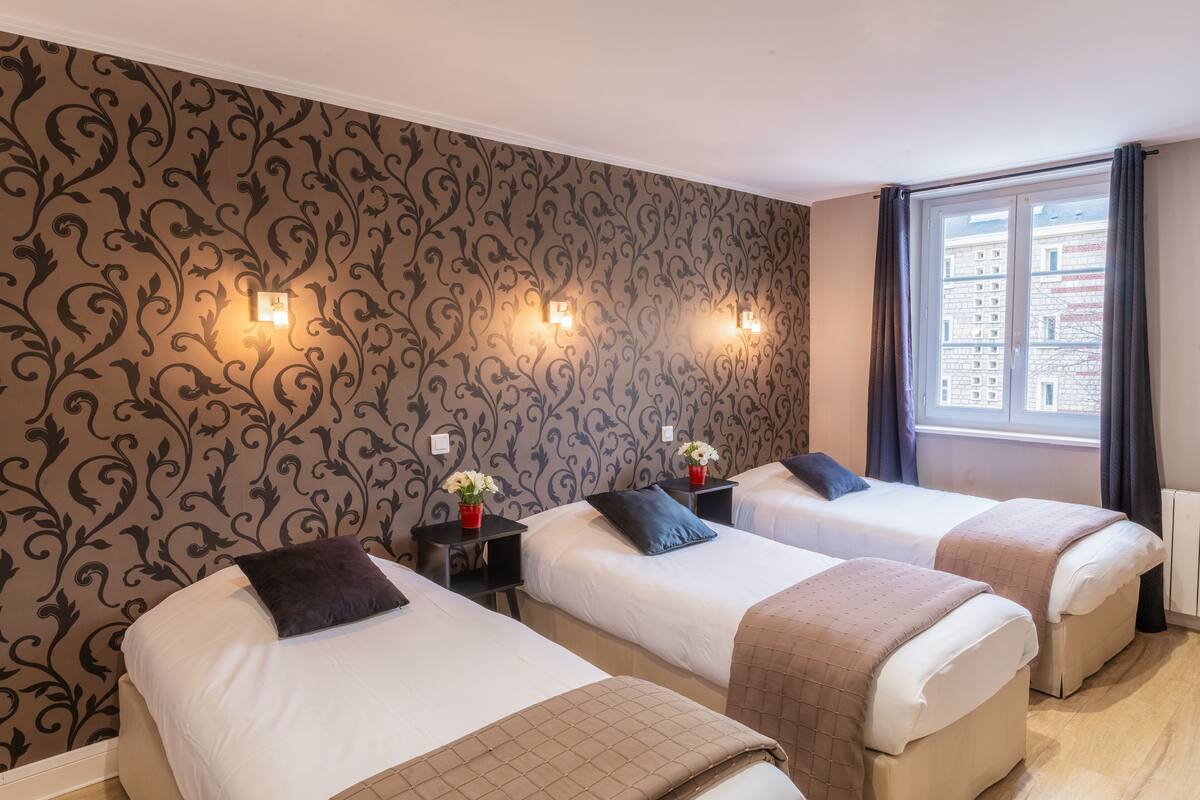 Hotelkamer met drie enkele bedden, behang met een patroon, twee wandlampen, een raam met gordijnen en uitzicht op gebouwen.