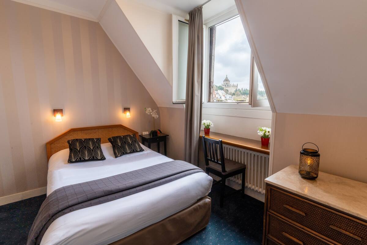 Een slaapkamer met een bed, nachtkastjes, een bureau, een stoel, een radiator, een raam met gordijnen en uitzicht op een stadsgezicht.