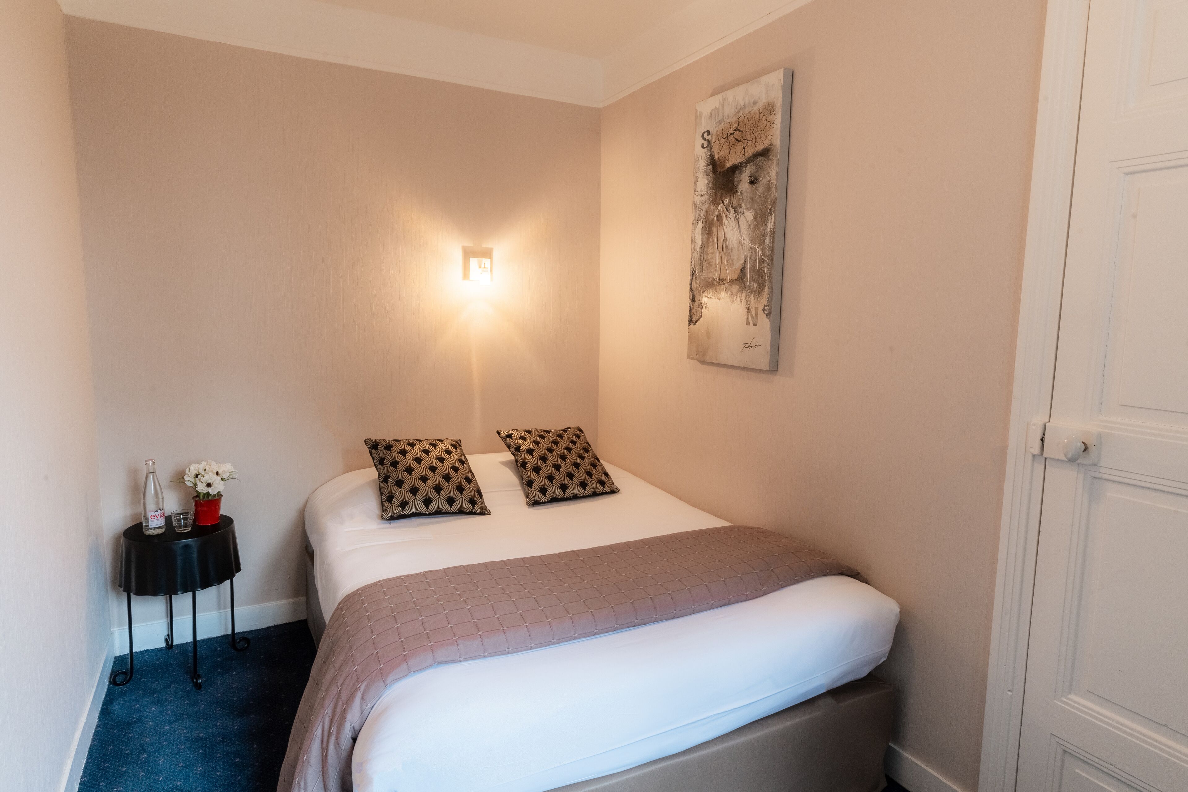 A neatly made bed with a white comforter and two patterned pillows, a bedside table with a bottle and flowers, and a wall-mounted light fixture.