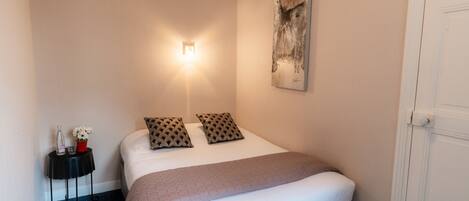 A neatly made bed with a white comforter and two patterned pillows, a bedside table with a bottle and flowers, and a wall-mounted light fixture.