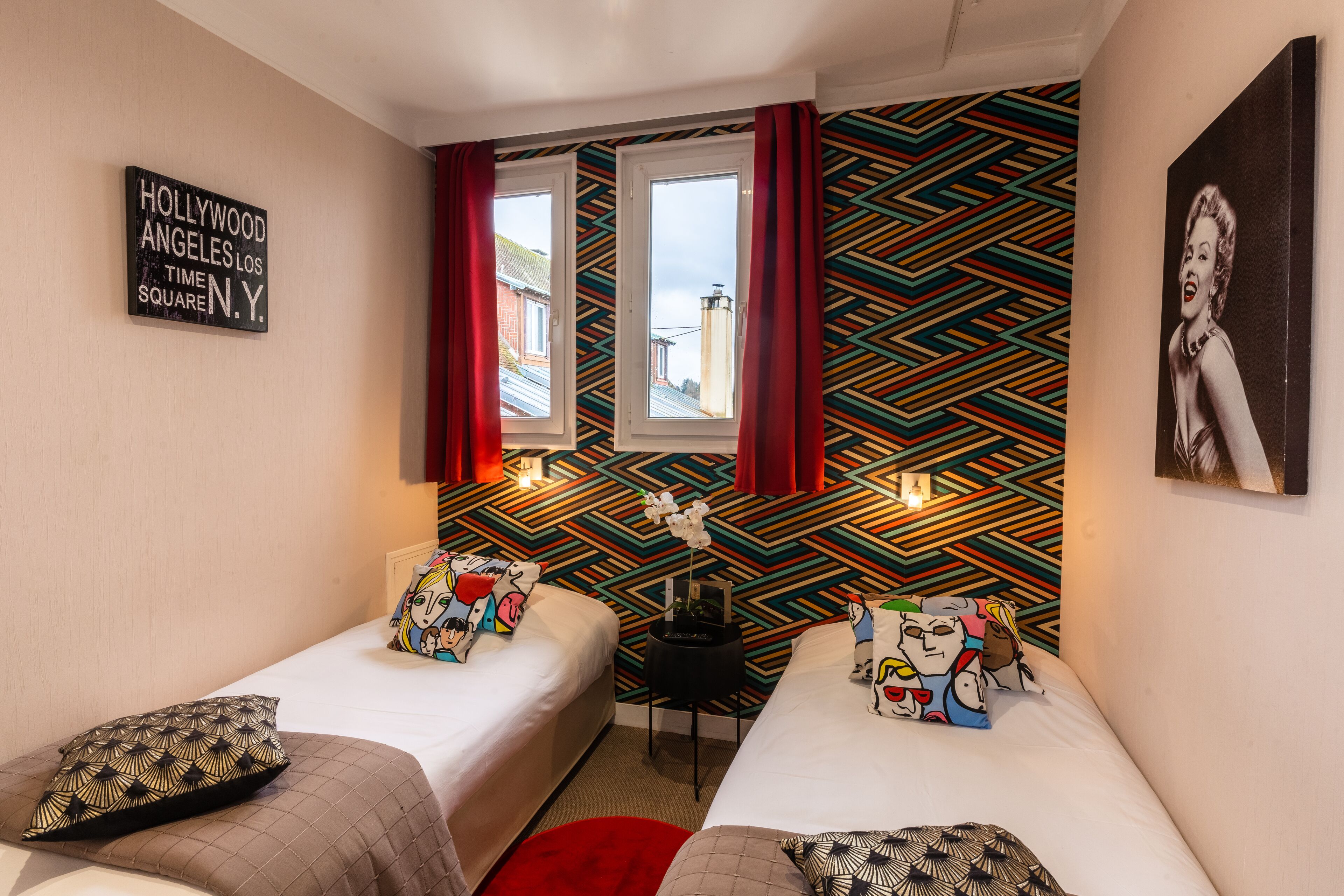 A hotel room with two beds, a colorful geometric wall pattern, a 'Hollywood Los Angeles Time Square N.Y.' sign, and a framed picture on the wall.