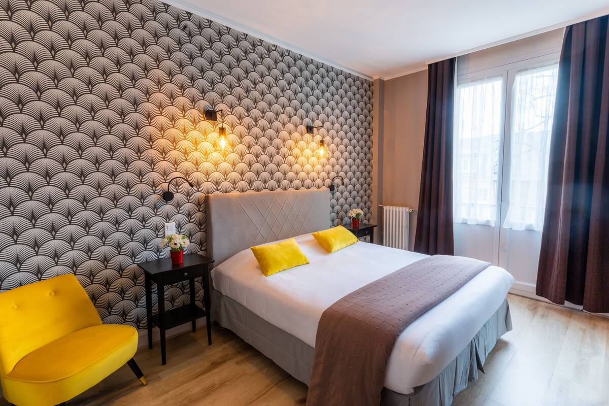 Een hotelkamer met behang met een patroon, een bed met twee kussens, een nachtkastje met een lamp, een stoel en een raam met gordijnen.