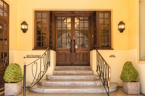 Une porte à deux battants en bois, ornée de panneaux de verre, flanquée de deux lanternes fixées au mur, qui mène à un escalier doté d’une rampe en métal.