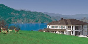 Una casa de dos pisos con balcón, cerca de un lago con montañas al fondo.