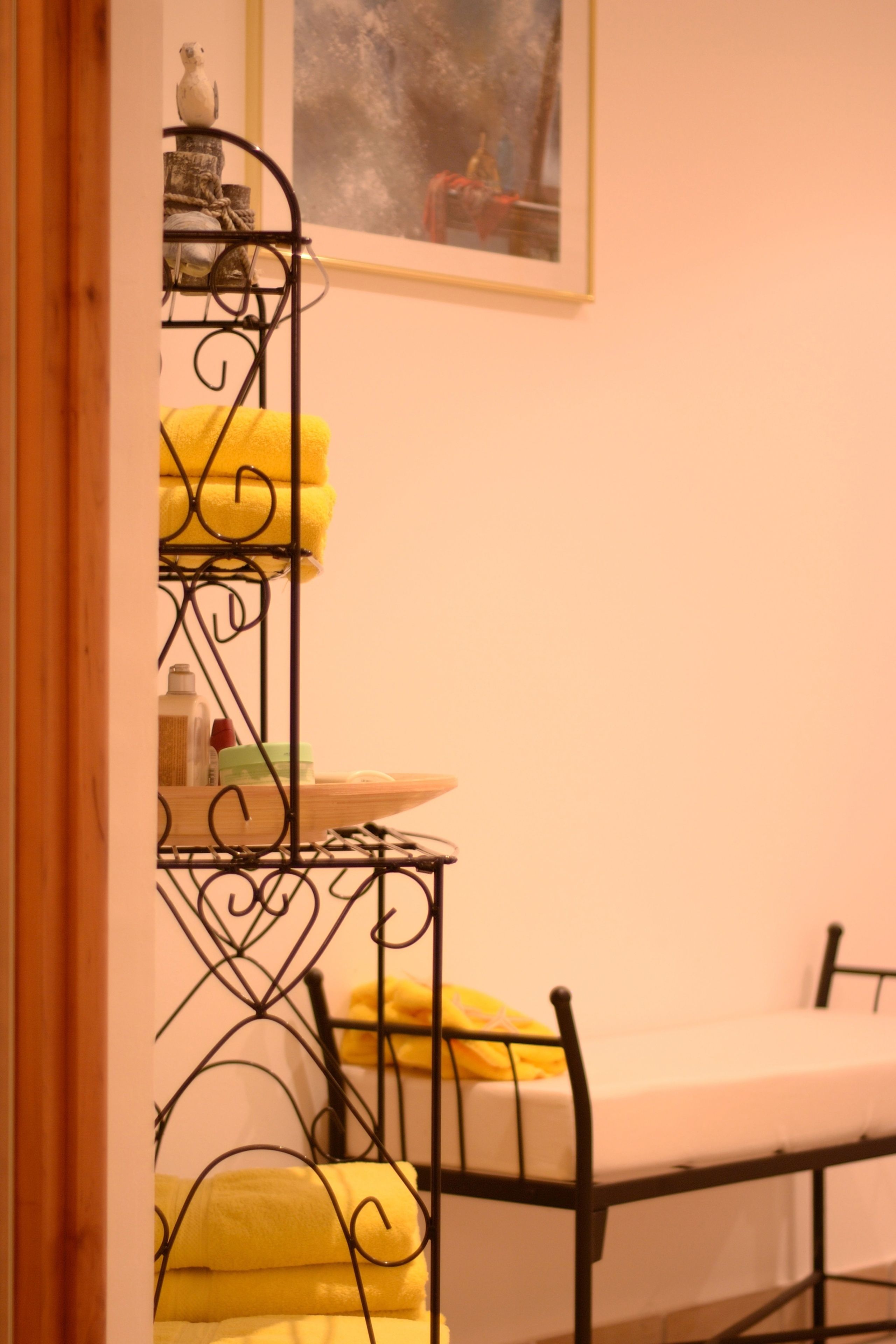 Een kamer met een metalen bedframe, een wit matras en gele handdoeken. Er hangt een schilderij aan de muur en een plank met verschillende spullen.