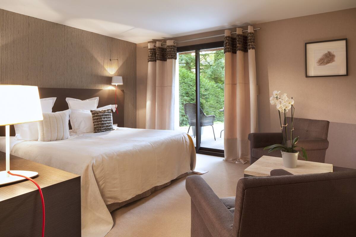 A hotel room with a large bed, a desk with a lamp, a chair, a potted plant, and a view of greenery through the window.