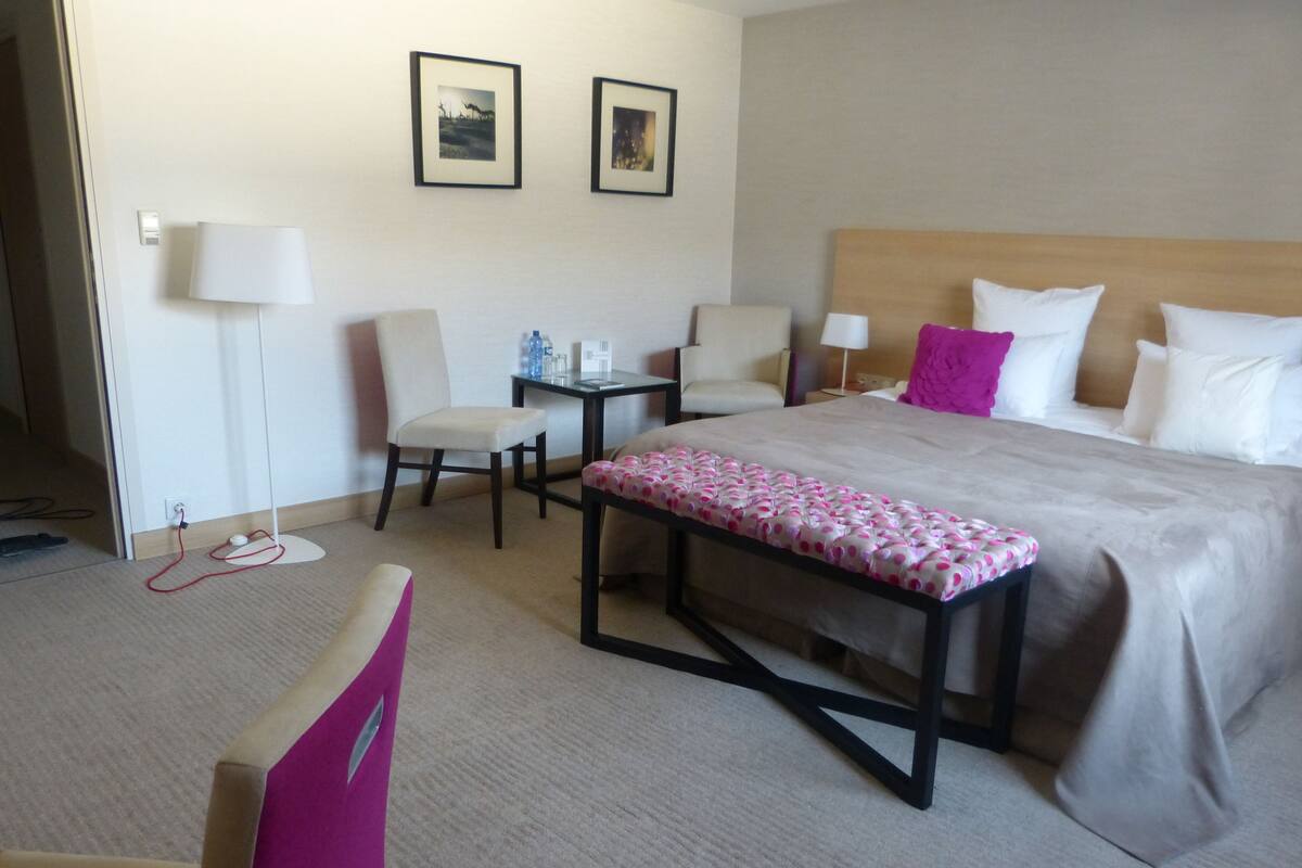 A hotel room with a bed, two chairs, a small table, a lamp, and a bench.