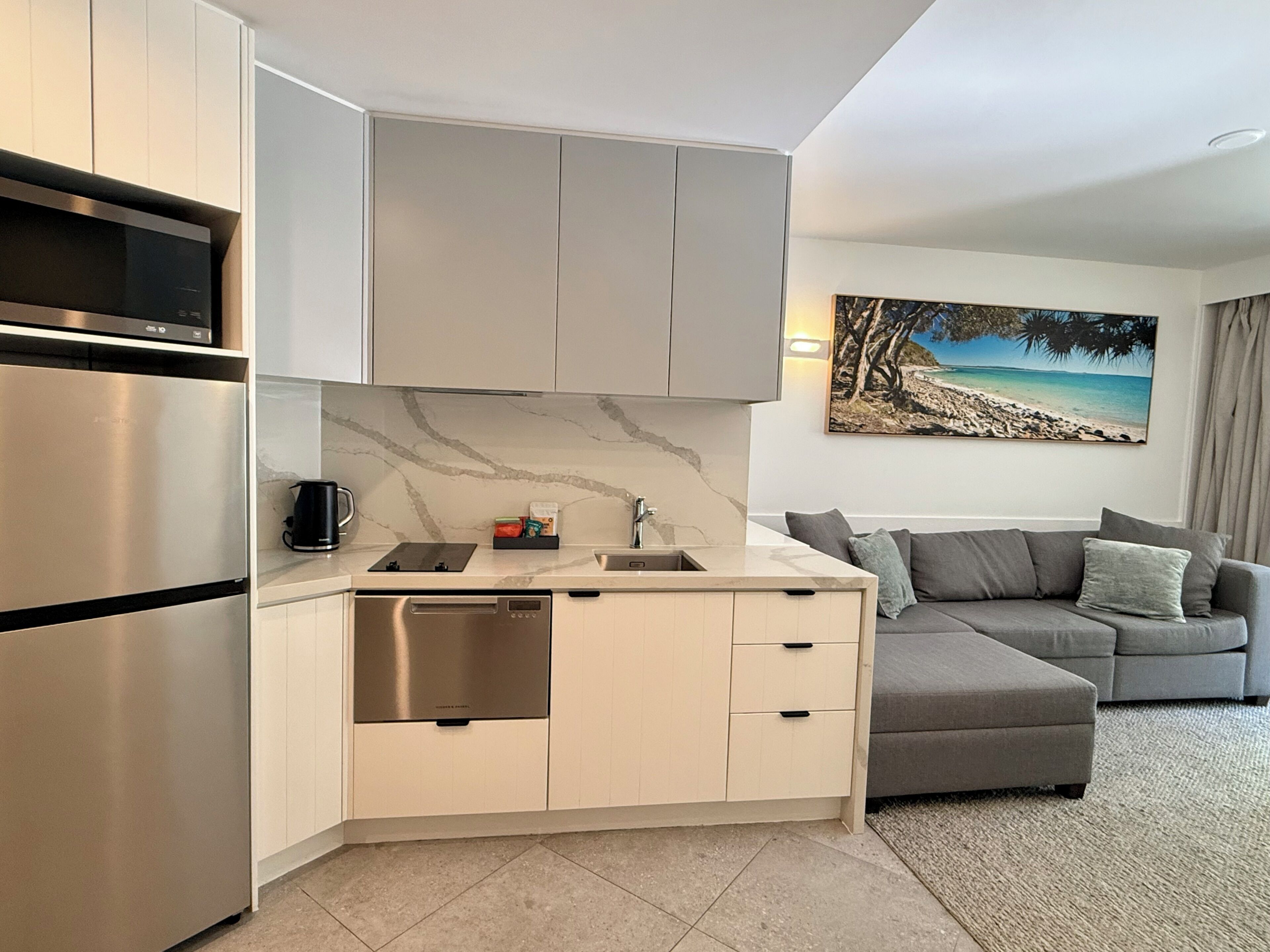 A modern kitchen with a built-in oven, microwave, and dishwasher. There is a gray sofa and a painting of a beach scene on the wall.