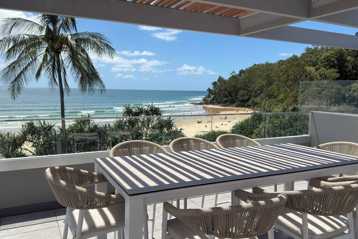 A balcony with a view of the beach, a table, and wicker chairs.