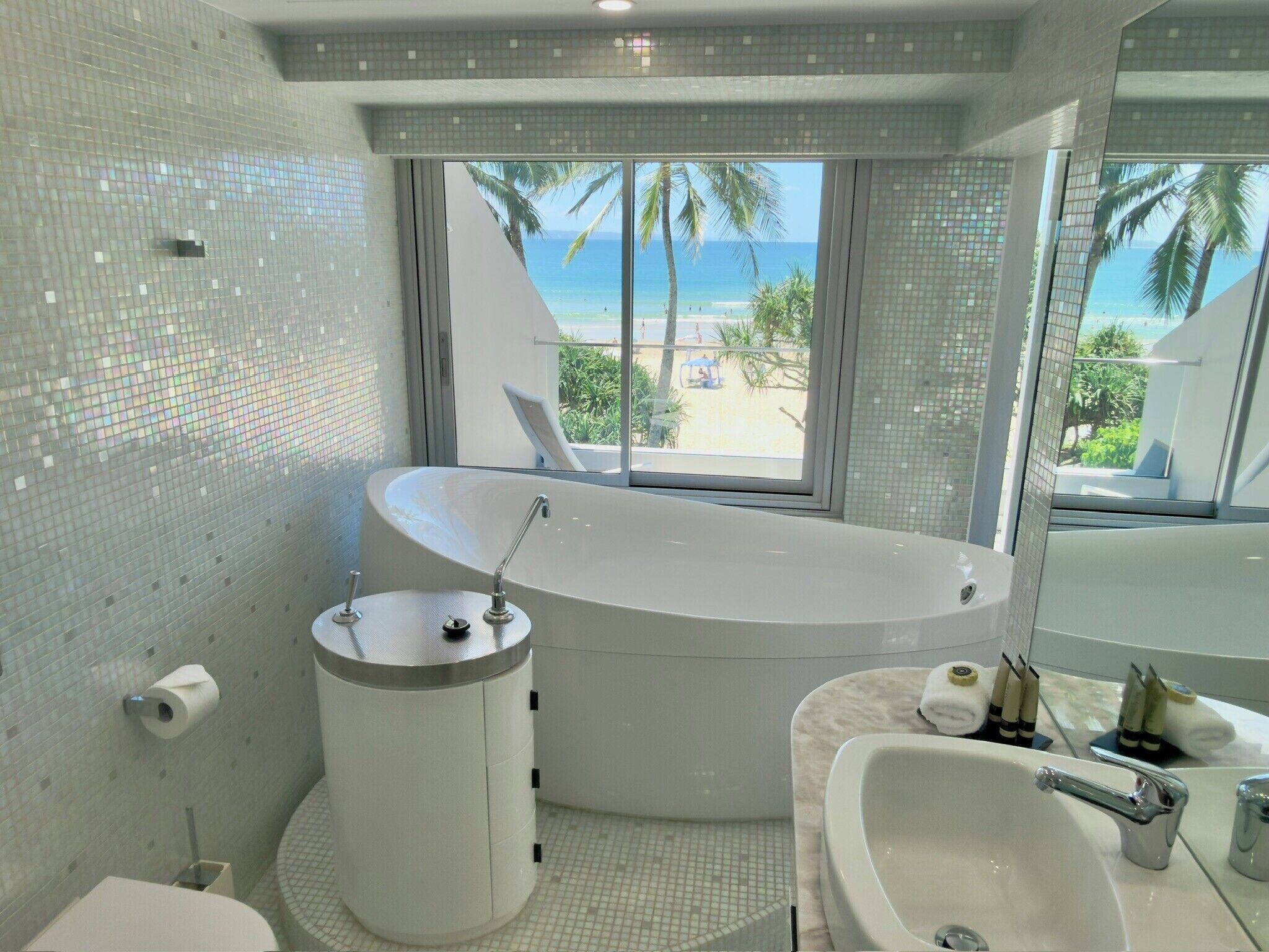 A bathroom with a large bathtub, a separate sink, and a toilet. There is a window providing a view of the beach and palm trees.