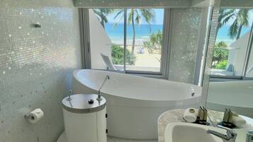 A bathroom with a large bathtub, a separate sink, and a toilet. There is a window providing a view of the beach and palm trees.