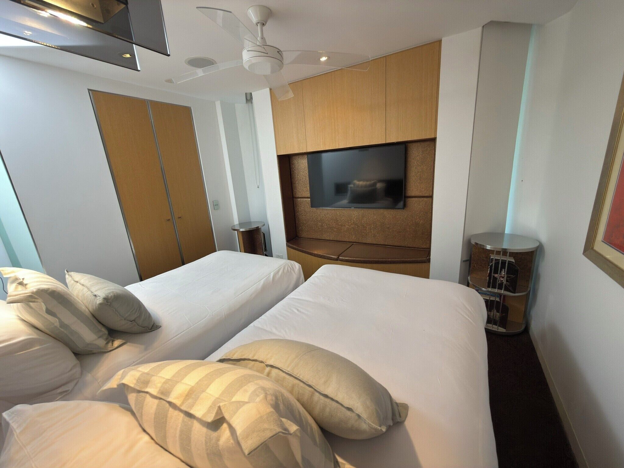 Habitación de hotel con dos camas, un ventilador de techo, un televisor montado en la pared y una mesita con una lámpara.