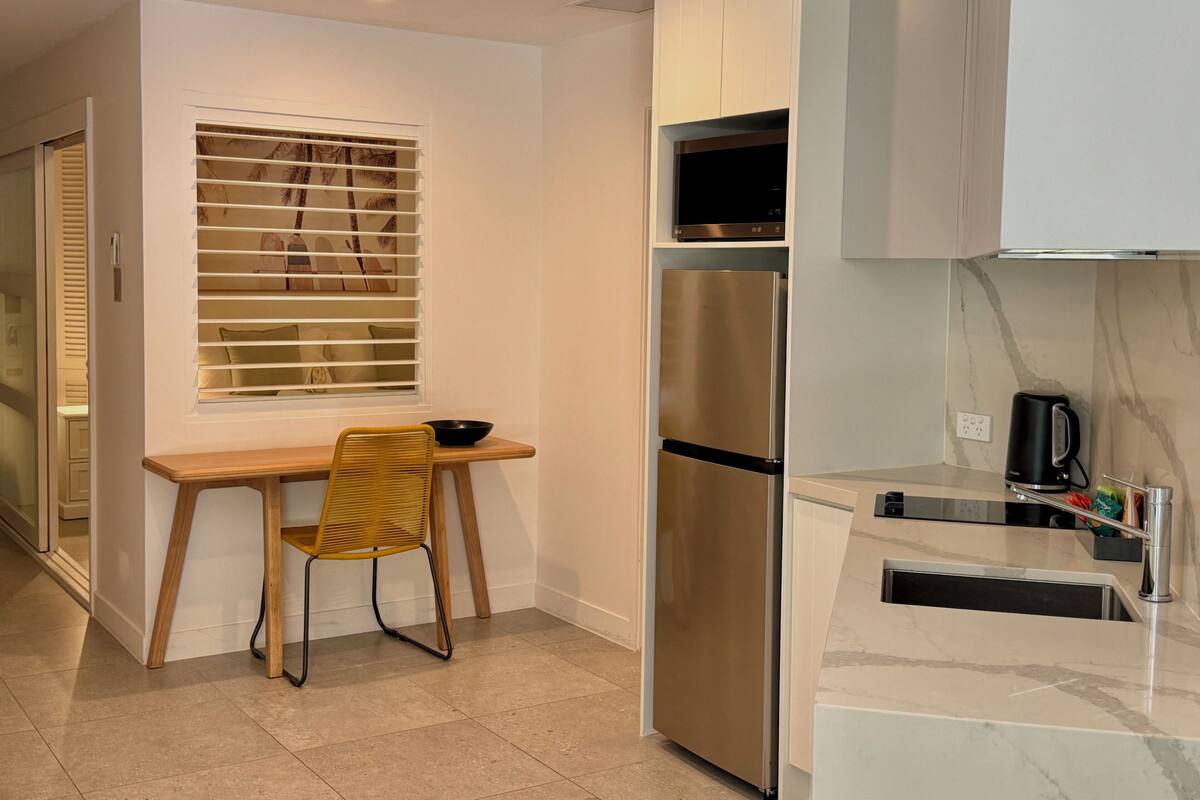 A compact kitchen with a built-in refrigerator, a wooden dining table with a chair, and a window with blinds.